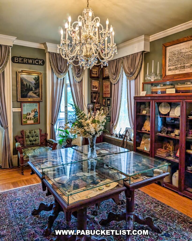 Elegant room at the Berwick Historical Society Museum in Columbia County, PA, featuring a crystal chandelier, Victorian-style furniture, framed historic maps and artwork, display cabinets filled with Berwick memorabilia, and a glass-topped table showcasing photographs and artifacts related to the town’s history.