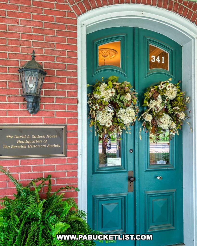 Front entrance of the historic David A. Sadock House in Berwick, Pennsylvania, featuring double teal doors adorned with floral wreaths, a brass plaque identifying it as the headquarters of the Berwick Historical Society, and a glimpse of the ornate chandelier inside through the glass panels.