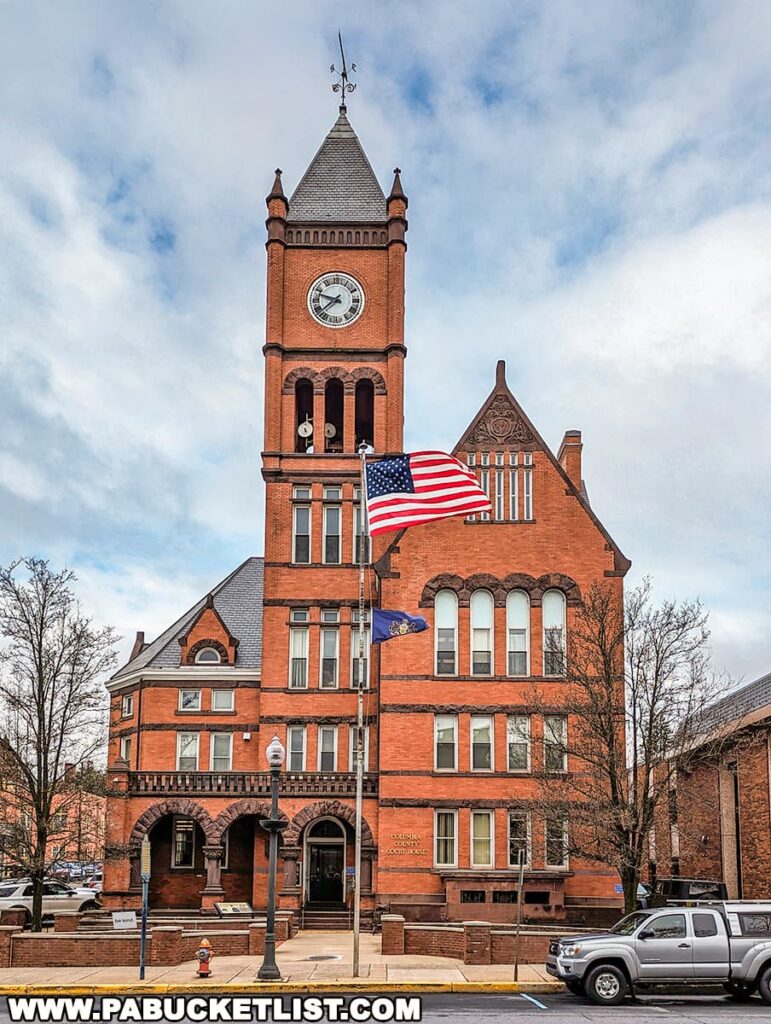 Historic Columbia County Courthouse in Bloomsburg, Pennsylvania, featuring a tall red-brick clock tower, arched entrance, and American and Pennsylvania state flags flying in front of the building.