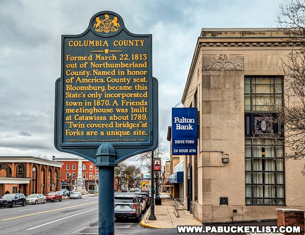 Columbia County historical marker in Bloomsburg, Pennsylvania, describing the county’s formation in 1813, its county seat, and notable historic sites, with downtown Bloomsburg buildings and traffic in the background.