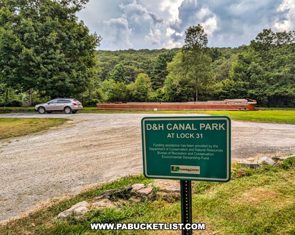 Parking area at the D\&H Canal Park at Lock 31 in Wayne County, Pennsylvania, with a green informational sign in the foreground and the life-sized replica canal boat visible in the background near the tree line.