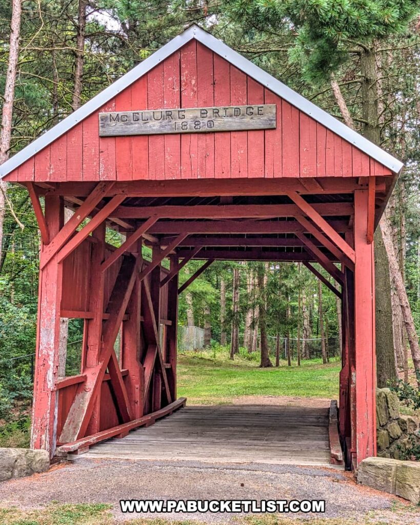 Front entrance view of the McClurg Covered Bridge in Washington County, Pennsylvania, showing its red wooden structure with a sign reading “McClurg Bridge 1880” above the opening and a grassy park landscape visible beyond.