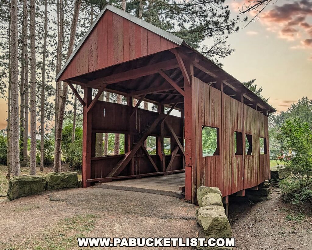 Angled view of the McClurg Covered Bridge in Washington County, Pennsylvania, a small red 1880 Kingpost truss bridge surrounded by tall trees at Hanover Township Park.
