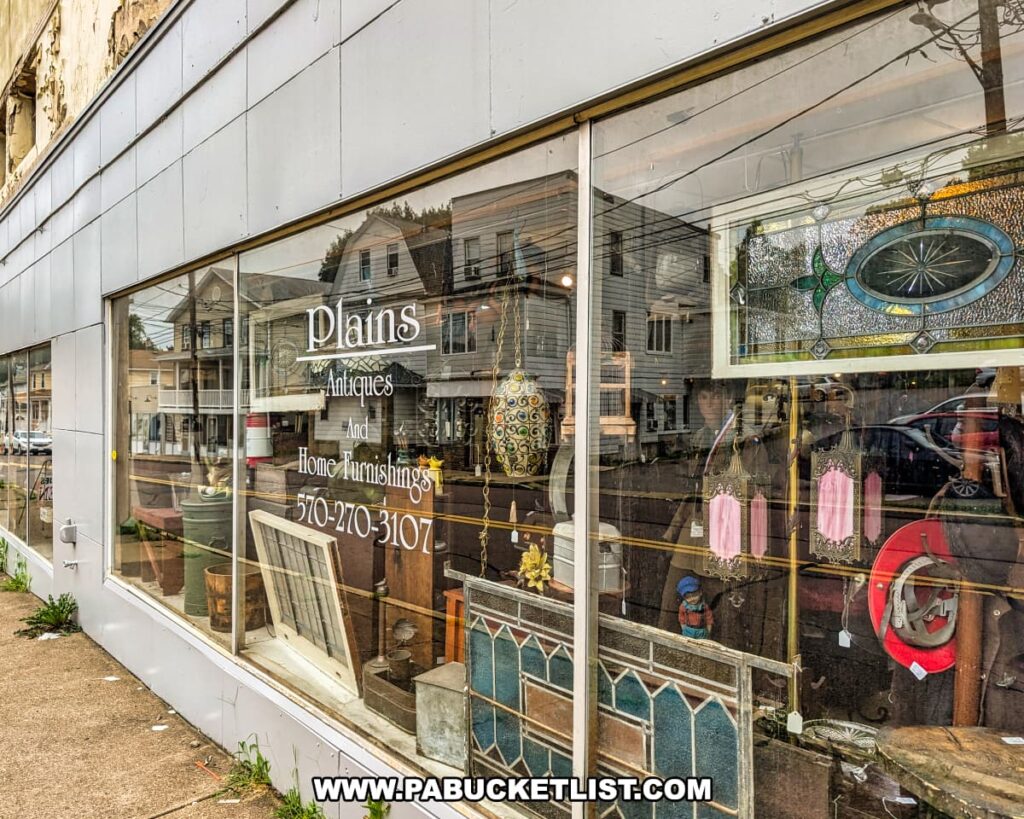 Storefront window of Plains Antiques and Home Furnishings in Luzerne County, PA, displaying vintage stained glass panels, lamps, and assorted antiques, with reflections of nearby buildings on the glass exterior of this two-level multi-vendor antique shop near Wilkes-Barre.
