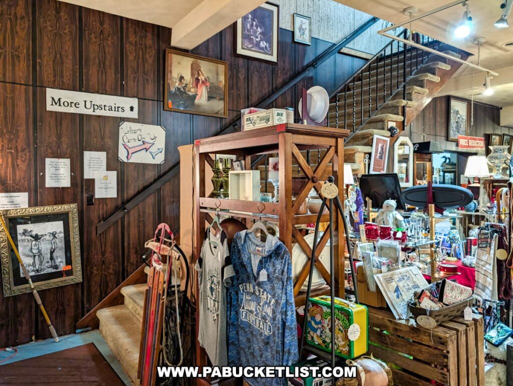 Staircase inside Plains Antiques in Luzerne County, PA, with a sign directing visitors upstairs, surrounded by vendor displays featuring vintage clothing, collectibles, framed artwork, glassware, and decorative items in this two-level multi-vendor antique shop near Wilkes-Barre.