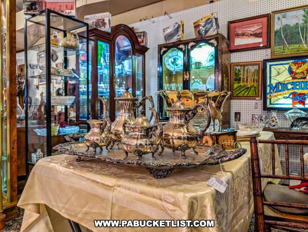 Ornate vintage silver tea set displayed on an embroidered tablecloth at Plains Antiques in Luzerne County, PA, surrounded by glass display cabinets, framed artwork, and stained glass in this two-level multi-vendor antique shop near Wilkes-Barre.