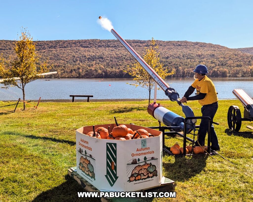 A man in a yellow shirt and helmet operates an air cannon launching a pumpkin high into the air over Sayers Lake during the Punkin Chunkin Fall Festival at Bald Eagle State Park in Centre County, PA, with a large bin of pumpkins in the foreground and autumn foliage covering the distant hills.