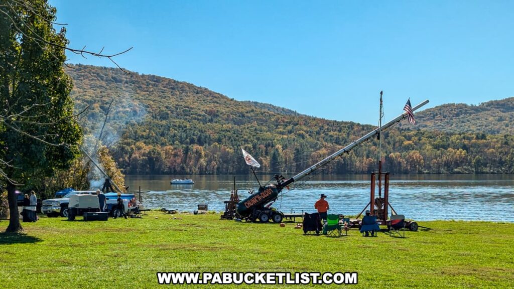 Spectators watch from lawn chairs as a large air cannon decorated with an American flag and a “Chunkinology" logo is set up along the shore of Sayers Lake during the Punkin Chunkin Fall Festival at Bald Eagle State Park in Centre County, PA, with wooded hillsides in peak autumn color rising in the background.