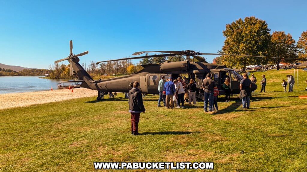 Visitors explore the interior of a military helicopter on display along the beach at Sayers Lake during the Punkin Chunkin Fall Festival at Bald Eagle State Park in Centre County, PA, with families gathered nearby and autumn foliage in the background.