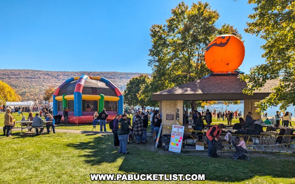 Families gather near a colorful inflatable bounce house and a pavilion topped with a giant inflatable pumpkin at the Punkin Chunkin Fall Festival at Bald Eagle State Park in Centre County, PA, with autumn foliage and Sayers Lake in the background.
