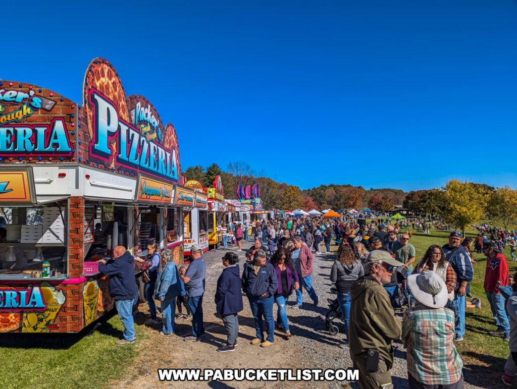 Large crowds line up at colorful food vendor trailers, including a pizzeria stand, during the Punkin Chunkin Fall Festival at Bald Eagle State Park in Centre County, PA, with additional vendor tents and autumn foliage in the background.