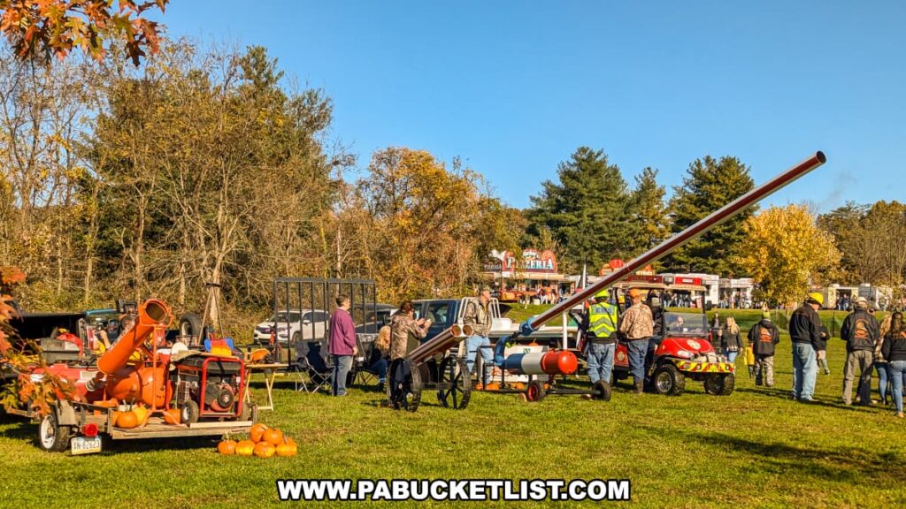 Festival participants gather around several air cannons and equipment trailers loaded with pumpkins at the Punkin Chunkin Fall Festival at Bald Eagle State Park in Centre County, PA, with food vendors and colorful autumn foliage in the background.