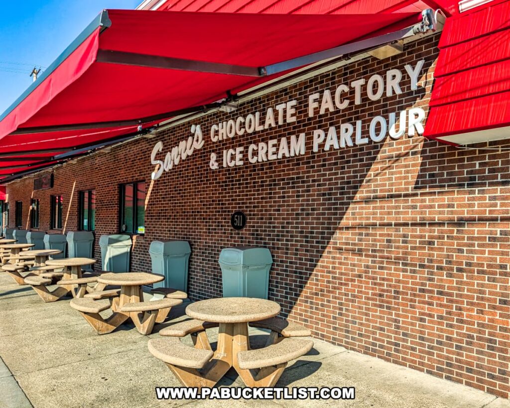 Outdoor seating area at Sarris Candies’ Chocolate Factory and Ice Cream Parlour in Canonsburg, PA, featuring round picnic tables under bright red awnings along the brick building with the shop’s name displayed on the wall.
