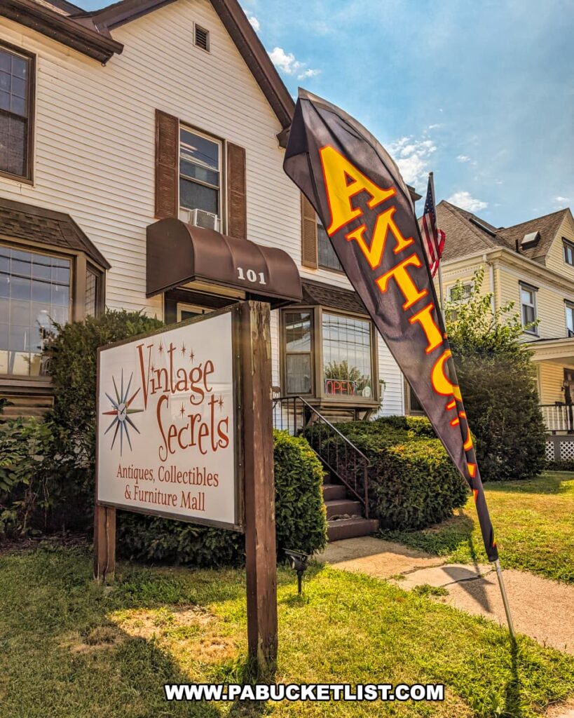 Exterior view of Vintage Secrets Antiques in Canonsburg, Washington County, PA, showing the shop’s sign reading “Antiques, Collectibles & Furniture Mall,” a tall black flag with bold orange “Antiques” lettering, and the three-story house that hosts the multi-vendor mall in the background.