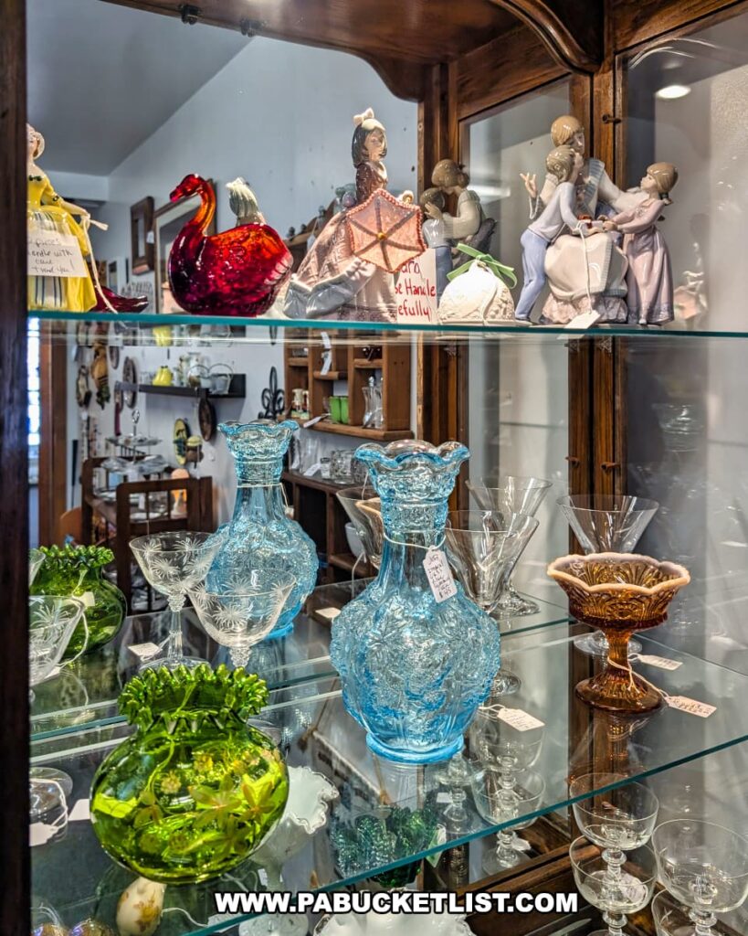 Glass display case at Vintage Secrets Antiques in Canonsburg, Washington County, PA, showcasing colorful vintage glassware including blue patterned vases, green and amber bowls, delicate stemware, and porcelain figurines in the multi-vendor antique mall.