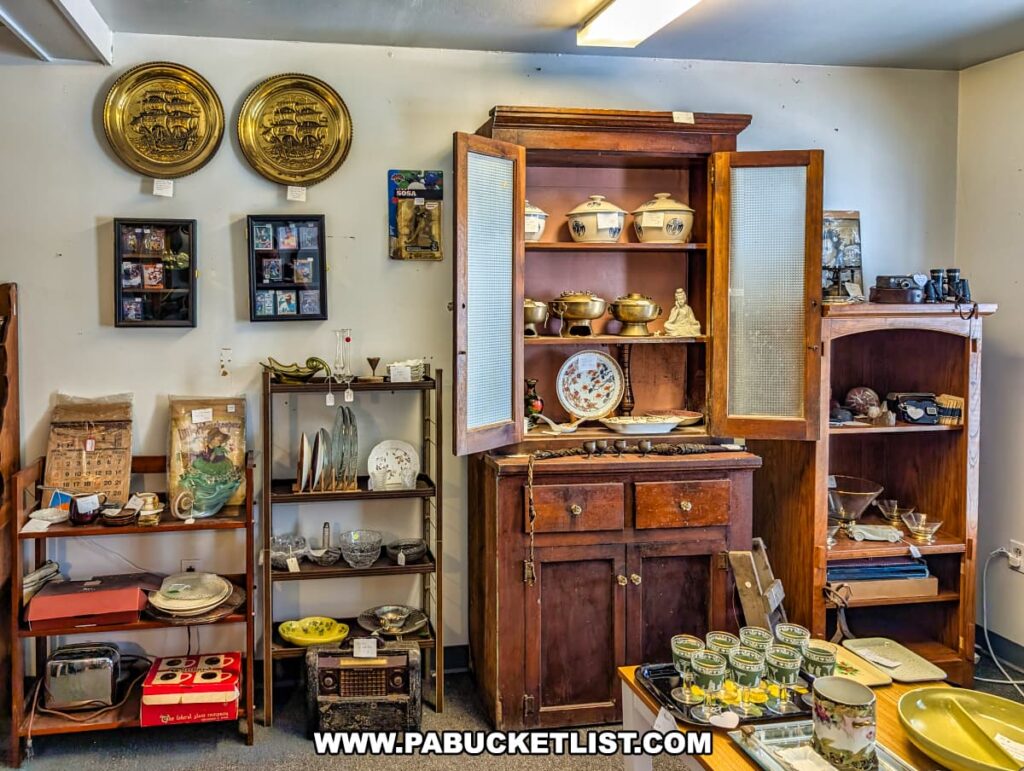 Vendor booth at Vintage Secrets Antiques in Canonsburg, Washington County, PA, showcasing vintage home décor including brass wall plates, framed art, glassware, ceramics, an antique wooden cabinet filled with collectibles, and assorted retro household items.