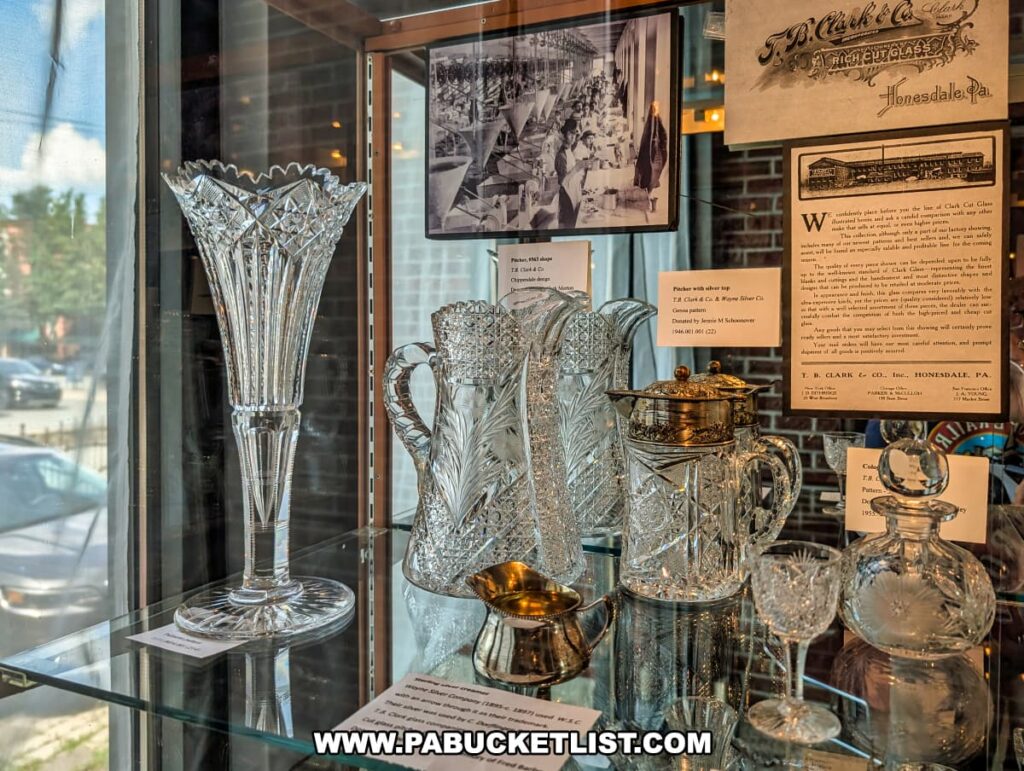 Display of ornate cut glassware produced by T.B. Clark & Co. in Honesdale, Pennsylvania, showcased at the Wayne County Historical Society Museum, with vintage advertisements and historic photos of the glassworks factory in the background.