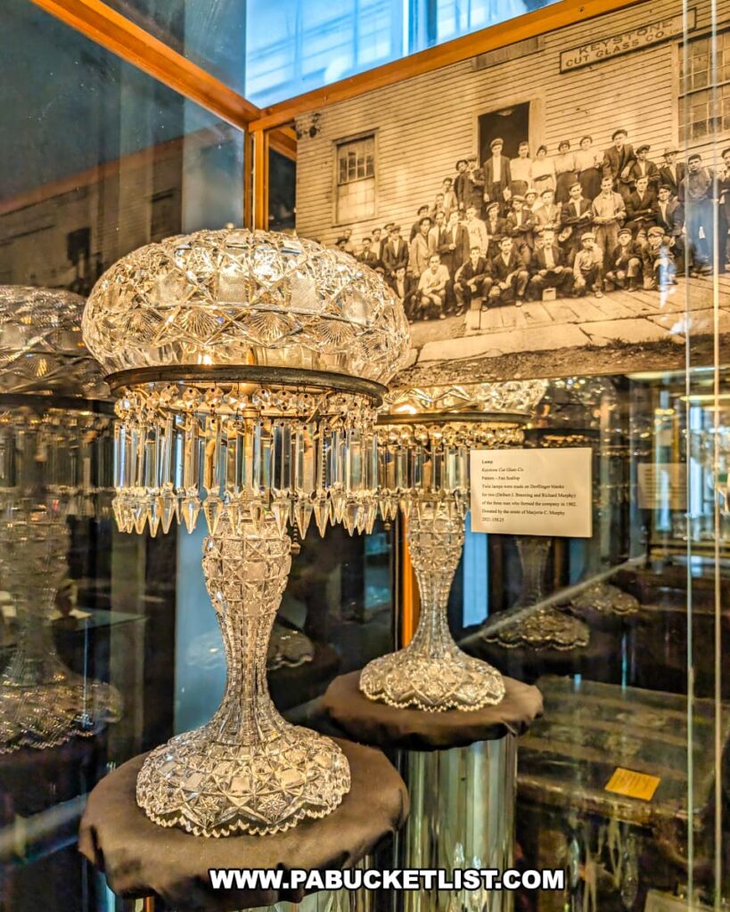 Intricately cut glass lamp made by the Keystone Cut Glass Company on display at the Wayne County Historical Society Museum in Honesdale, Pennsylvania, with a historic black-and-white photo of factory workers in the background.
