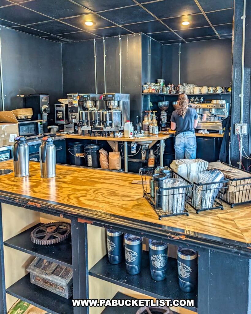 Interior of Black and Brass Coffee Roasting Company near Carley Brook Falls in Wayne County, PA, showing a barista preparing drinks behind a wooden counter with espresso machines, coffee equipment, and shelves of mugs and supplies in a cozy industrial-style setting.