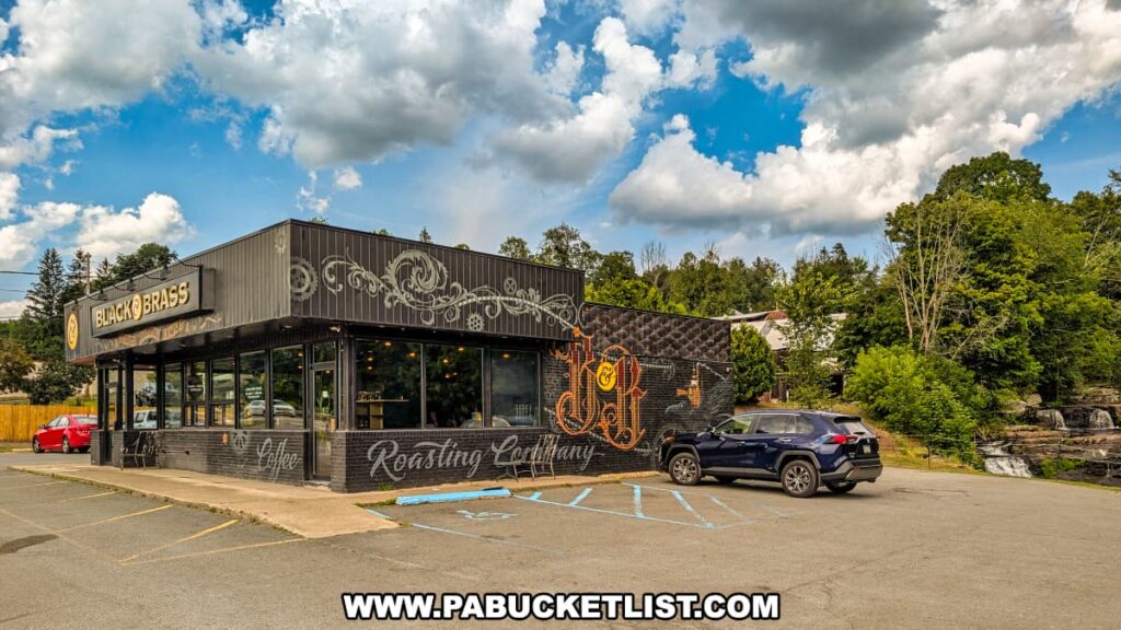 Exterior view of Black and Brass Coffee Roasting Company along Route 6 near Honesdale, PA, featuring the café’s black brick building with decorative gold lettering and murals, cars parked in front, and the scenic Carley Brook Falls visible beside the shop.