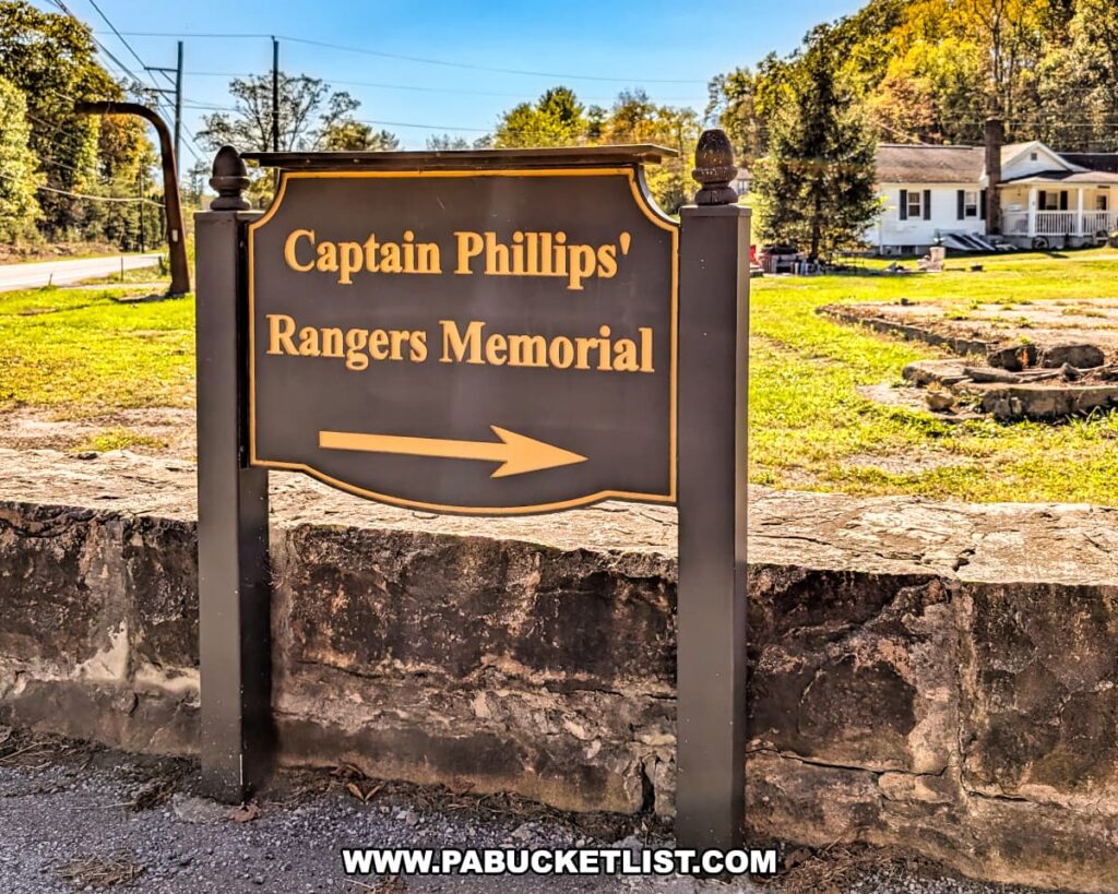 Directional sign along Route 26 in Bedford County pointing toward the Captain Phillips’ Rangers Memorial, with a stone wall, grassy lawn, and houses visible in the background under bright autumn sunlight.