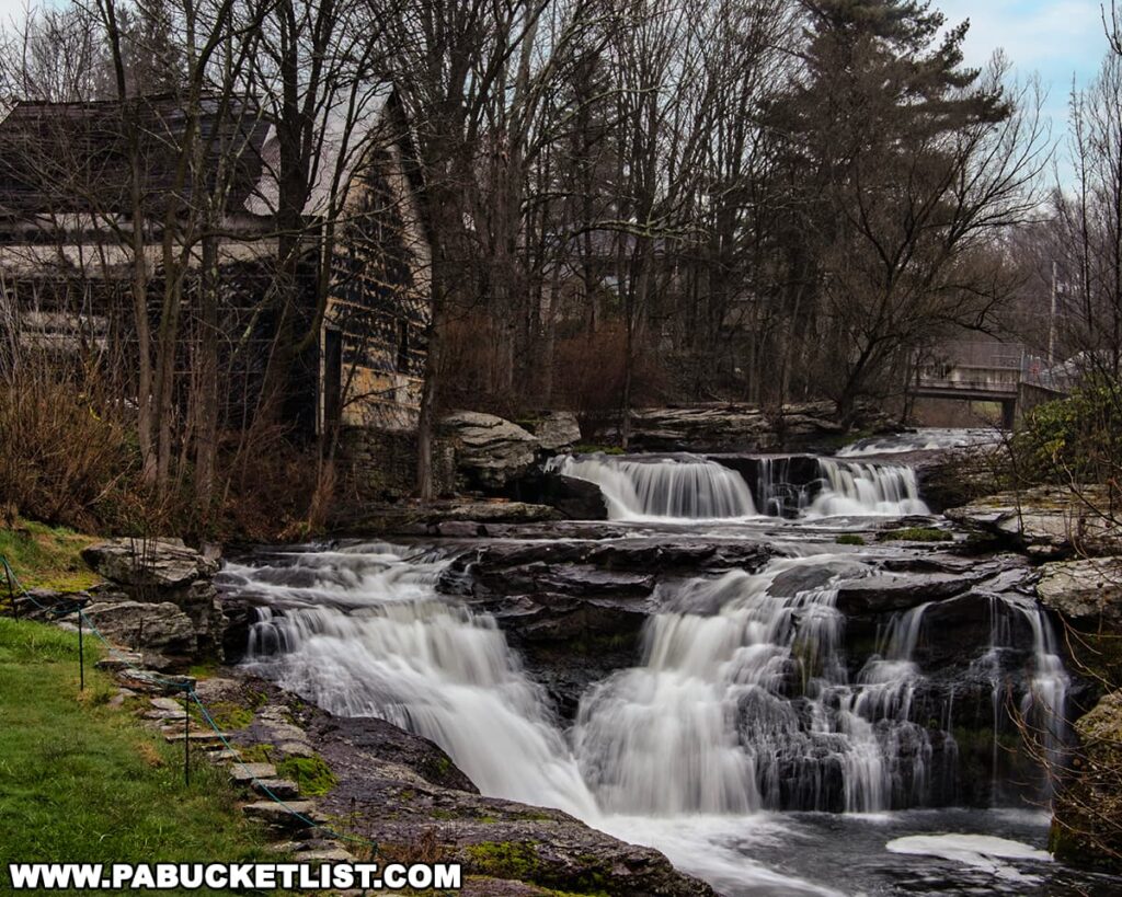 Carley Brook Falls in Wayne County, PA, a beautiful 20-foot multi-tiered waterfall cascading over dark rock ledges beside a weathered wooden building and surrounded by leafless early spring trees near Black and Brass Coffee Roasting Company along Route 6 near Honesdale.