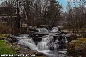Carley Brook Falls in Wayne County, PA, a 20-foot-tall multi-tiered waterfall cascading over dark rock ledges surrounded by trees, with an old weathered building and small bridge visible near the top of the falls along Route 6 near Honesdale.