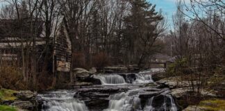 Carley Brook Falls in Wayne County, PA, a 20-foot-tall multi-tiered waterfall cascading over dark rock ledges surrounded by trees, with an old weathered building and small bridge visible near the top of the falls along Route 6 near Honesdale.