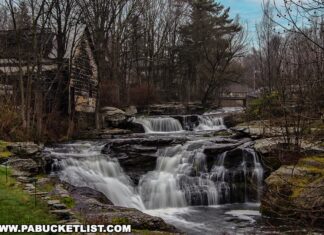 Carley Brook Falls in Wayne County, PA, a 20-foot-tall multi-tiered waterfall cascading over dark rock ledges surrounded by trees, with an old weathered building and small bridge visible near the top of the falls along Route 6 near Honesdale.