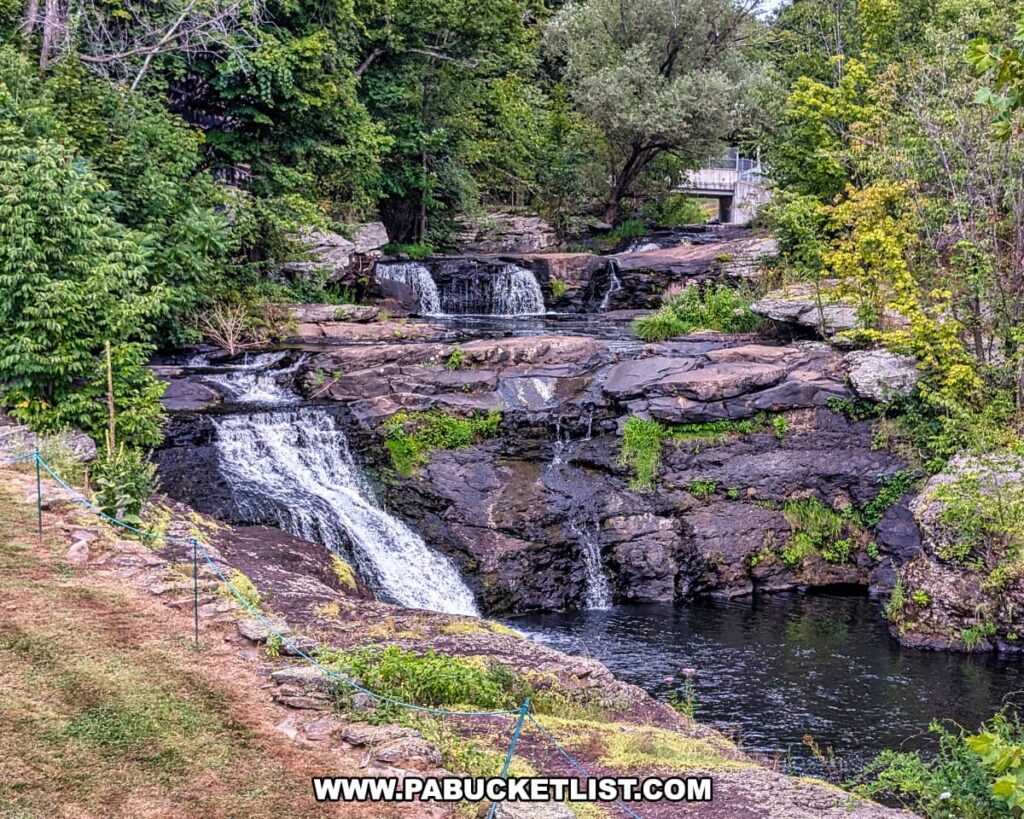 Carley Brook Falls in Wayne County, PA, a 20-foot multi-tiered waterfall flowing over dark rock ledges surrounded by lush summer greenery, with clear pools and a small bridge visible near the top of the falls along Route 6 near Honesdale.