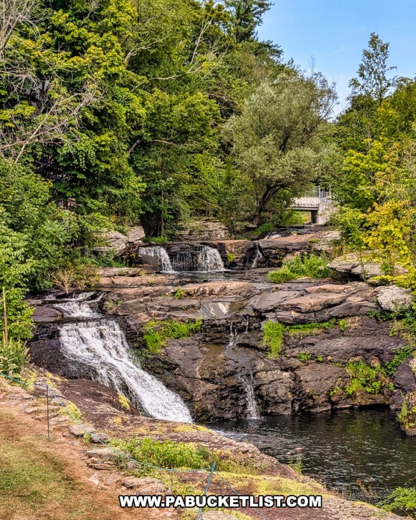 Carley Brook Falls in Wayne County, PA, a picturesque 20-foot multi-tiered waterfall cascading over layered rock formations surrounded by vibrant summer foliage, with a small bridge visible above the falls near Route 6 and Black and Brass Coffee Roasting Company in Honesdale.