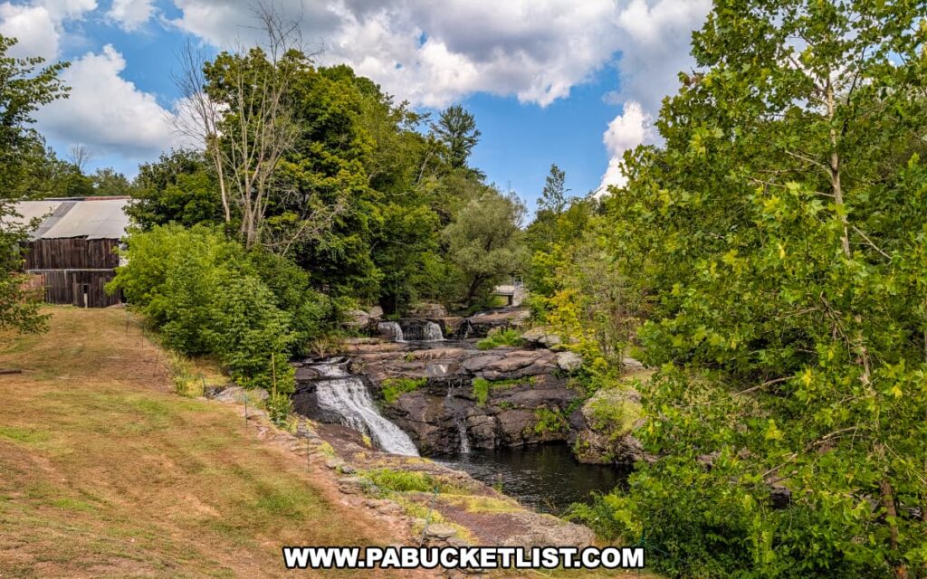 Carley Brook Falls in Wayne County, PA, a scenic 20-foot multi-tiered waterfall cascading over rock ledges into a calm pool, surrounded by lush green summer foliage and an old wooden building visible on the left near Route 6 and Black and Brass Coffee Roasting Company in Honesdale.