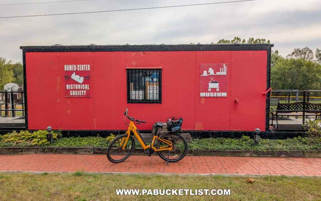 A bright yellow bicycle is parked in front of a red railcar that serves as the Homer-Center Historical Society Museum along the Hoodlebug Trail in Indiana County, Pennsylvania. The railcar features signs identifying it as a museum with graphics depicting a coal car and industrial buildings. A brick walkway and small garden border the railcar, while trees and park benches can be seen in the background. The museum sits beside the 10-mile Hoodlebug Trail, which follows the former route of the Pennsylvania Railroad’s Indiana Branch, once traveled by the “Hoodlebug” passenger coach.