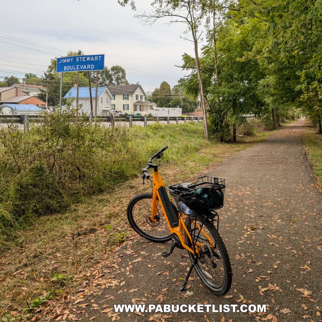 A bright yellow bicycle rests on the paved Hoodlebug Trail in Indiana County, Pennsylvania, near a blue roadside sign marking Jimmy Stewart Boulevard. The trail runs parallel to a row of houses and businesses in the distance, while trees line the opposite side, arching over the peaceful path scattered with fallen leaves. This section of the 10-mile rail trail follows the former route of the Pennsylvania Railroad’s Indiana Branch, once traveled by the self-propelled “Hoodlebug” passenger coach.