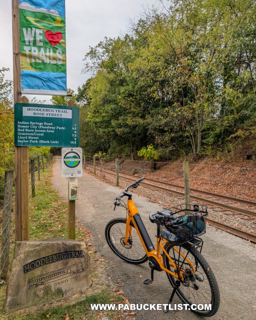 A bright yellow bicycle is parked at the Rose Street trailhead of the Hoodlebug Trail in Indiana County, Pennsylvania. Signs mark distances to various destinations along the 10-mile route, and a colorful banner reading “We ? Trails” flutters above the path. A large engraved stone identifies the trail beside the paved entrance, which runs parallel to railroad tracks bordered by autumn trees. The Hoodlebug Trail follows the former Pennsylvania Railroad’s Indiana Branch, once used by the self-propelled “Hoodlebug” passenger coach.