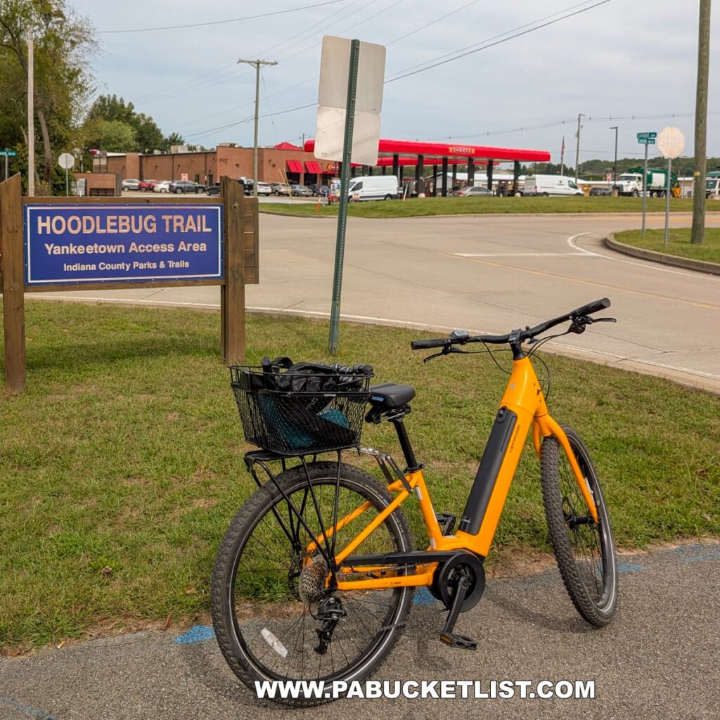 A bright yellow bicycle is parked near a wooden sign marking the Hoodlebug Trail Yankeetown Access Area in Indiana County, Pennsylvania. In the background, a busy intersection and a red-roofed Sheetz convenience store are visible under an overcast sky. This trailhead serves as one of several access points to the 10-mile Hoodlebug Trail, which follows the former Pennsylvania Railroad’s Indiana Branch once traveled by the self-propelled “Hoodlebug” passenger coach.