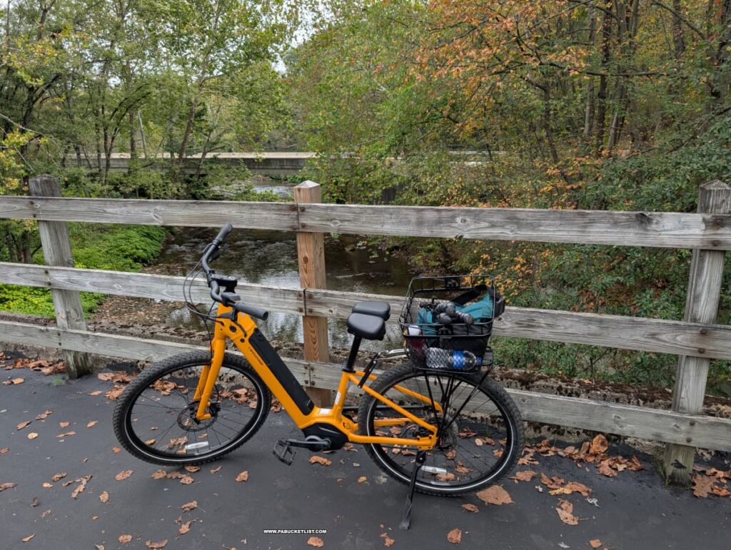A bright yellow bicycle with a black basket rests on a paved section of the Hoodlebug Trail in Indiana County, Pennsylvania, beside a wooden fence overlooking a gently flowing creek lined with early autumn foliage. Fallen leaves scatter across the trail, and a bridge is visible in the background among the trees. The peaceful scene captures the natural beauty along the 10-mile rail trail that follows the former route of the Pennsylvania Railroad’s Indiana Branch, once traveled by the self-propelled “Hoodlebug” passenger coach.