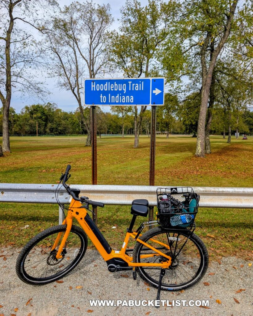 A bright yellow bicycle is parked beside a metal guardrail near a blue road sign reading “Hoodlebug Trail to Indiana” at Saylor Park in Indiana County, Pennsylvania. Fallen leaves scatter across the gravel shoulder, and trees with early autumn foliage frame the open grassy area in the background. This location marks the southern terminus of the 10-mile Hoodlebug Trail, which follows the former Pennsylvania Railroad’s Indiana Branch once traveled by the self-propelled “Hoodlebug” passenger coach.