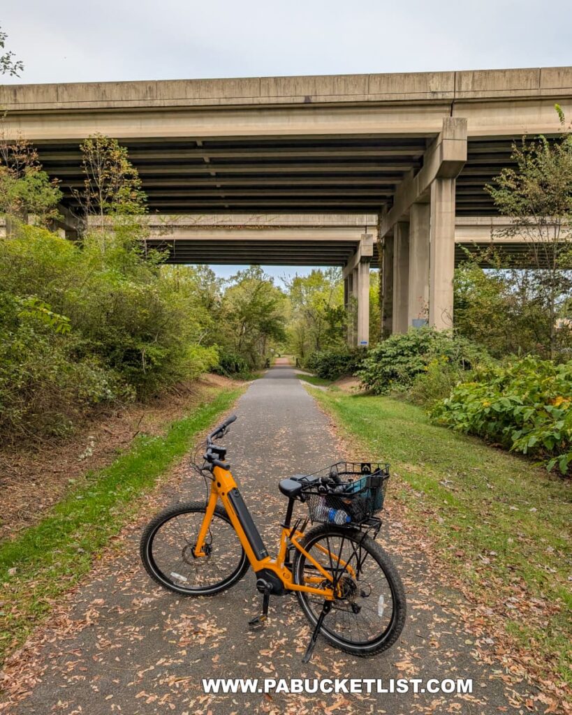 A bright yellow bicycle is parked on the paved Hoodlebug Trail beneath the towering concrete overpasses of Route 422 in Indiana County, Pennsylvania. The trail, lined with green vegetation and scattered autumn leaves, stretches straight ahead through the shaded corridor formed by the highway above. This section of the 10-mile rail trail follows the former Pennsylvania Railroad’s Indiana Branch, once traveled by the self-propelled “Hoodlebug” passenger coach.