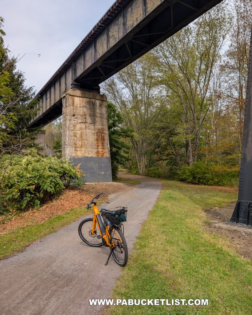 A bright yellow bicycle is parked on the paved Hoodlebug Trail beneath a towering steel railroad bridge supported by massive concrete piers in Indiana County, Pennsylvania. The trail curves gently through a wooded area with early autumn foliage, while the bridge spans high overhead, casting shadows across the path. This scenic section of the 10-mile rail trail follows the former Pennsylvania Railroad’s Indiana Branch, once used by the self-propelled “Hoodlebug” passenger coach.