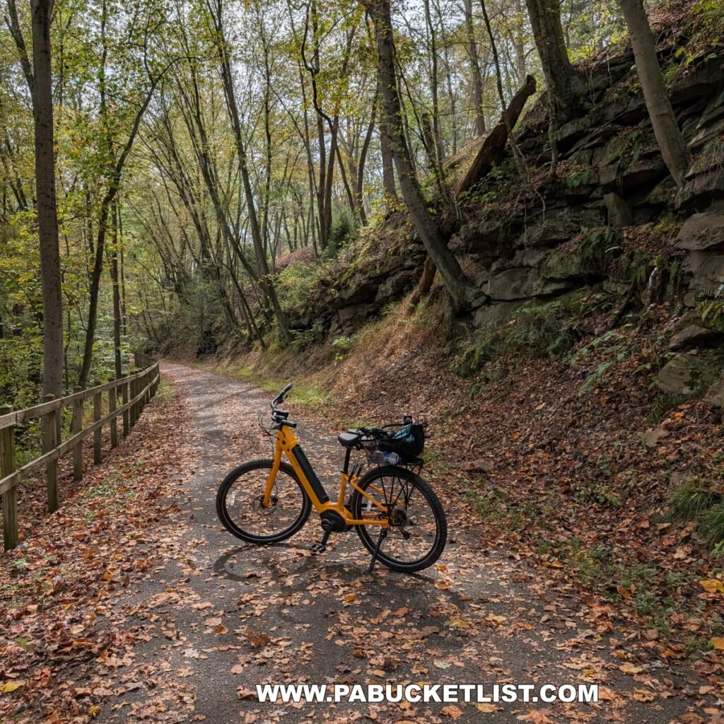 A bright yellow bicycle rests on a leaf-covered stretch of the Hoodlebug Trail that winds through a rocky, tree-lined corridor in Indiana County, Pennsylvania. A wooden fence borders one side of the paved path, while moss-covered rock ledges rise along the other. Dappled sunlight filters through the canopy of early autumn leaves, highlighting the serene, natural beauty of this section of the 10-mile rail trail that follows the former Pennsylvania Railroad’s Indiana Branch, once traveled by the self-propelled “Hoodlebug” passenger coach.
