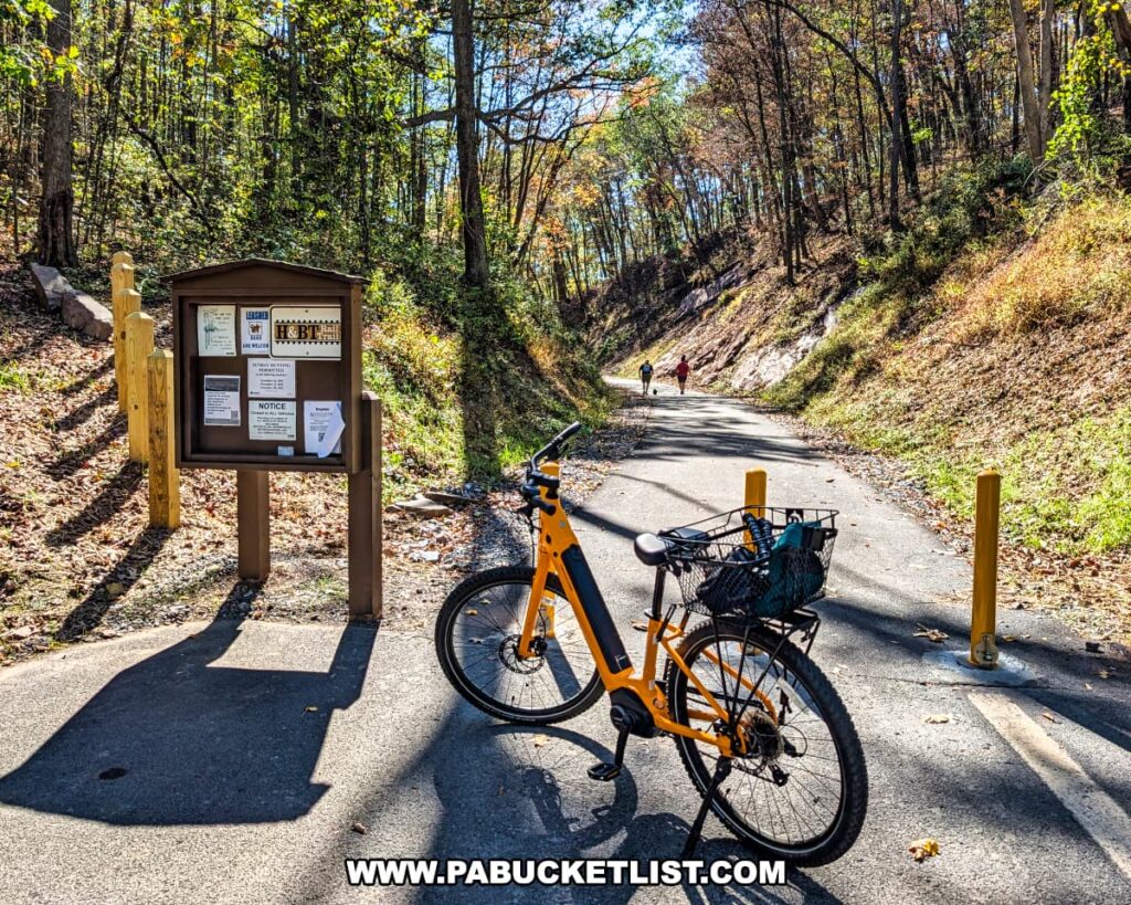A bright yellow bicycle with a basket is parked near an informational kiosk along the paved Huntingdon and Broad Top Rail Trail in Bedford County, Pennsylvania, surrounded by autumn foliage and sunlight filtering through the trees as two hikers walk along the trail in the distance.