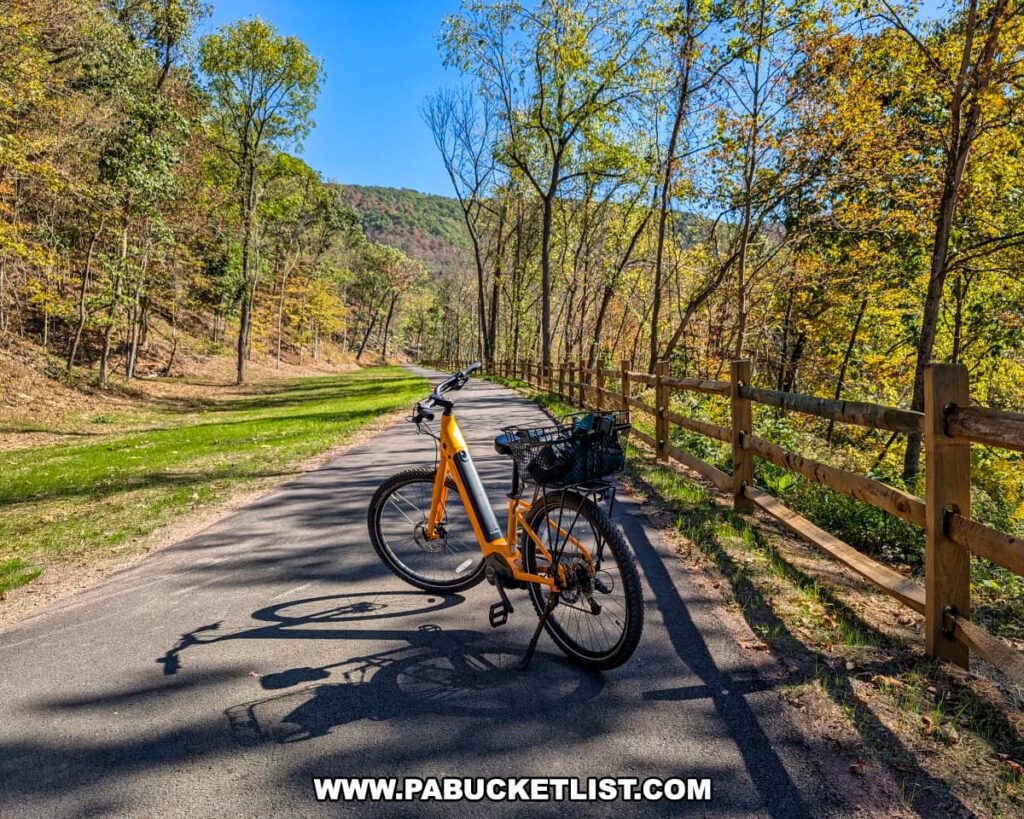 A yellow bicycle with a basket is parked on the paved Huntingdon and Broad Top Rail Trail in Bedford County, Pennsylvania, surrounded by early autumn foliage, wooden fencing, and rolling forested hills under a bright blue sky.