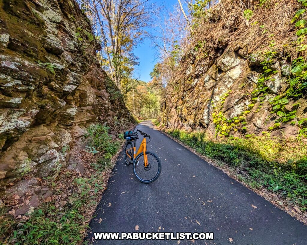 A yellow bicycle is parked on the paved Huntingdon and Broad Top Rail Trail in Bedford County, Pennsylvania, flanked by steep rock walls covered in moss and vines, with trees and blue sky visible beyond the narrow rocky passage.