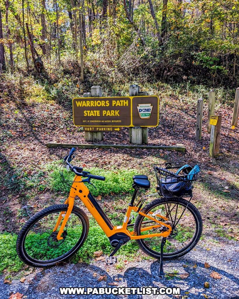 A bright yellow bicycle is parked near the entrance sign for Warriors Path State Park in Bedford County, Pennsylvania, marking the northern terminus of the Huntingdon and Broad Top Rail Trail on a sunny autumn day.