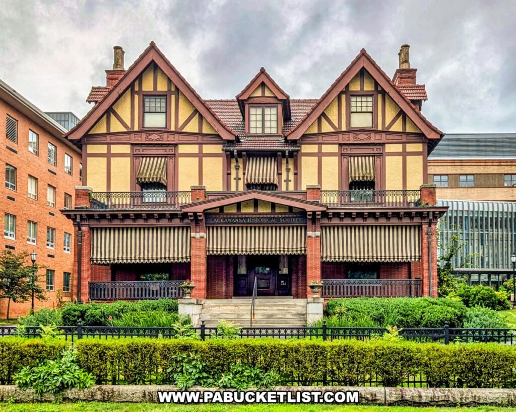 Front view of the historic Catlin House in Scranton, Pennsylvania, home to the Lackawanna Historical Society Museum. The Tudor Revival–style mansion features decorative half-timbering, striped awnings, red brick accents, and a covered front porch framed by lush greenery and an iron fence. The museum preserves and showcases the rich history and culture of Scranton and Lackawanna County.