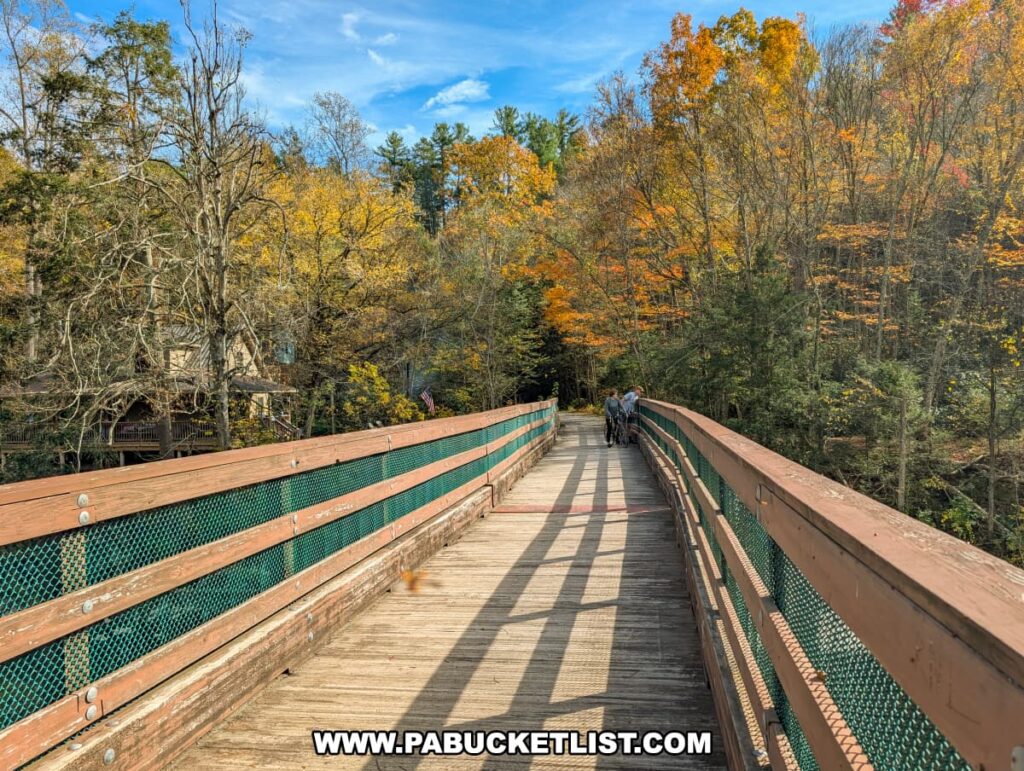 A wooden pedestrian bridge with green railings spans Penn’s Creek along the Penn’s Creek Path in Bald Eagle State Forest, surrounded by colorful autumn trees and blue skies as two cyclists cross in the distance.