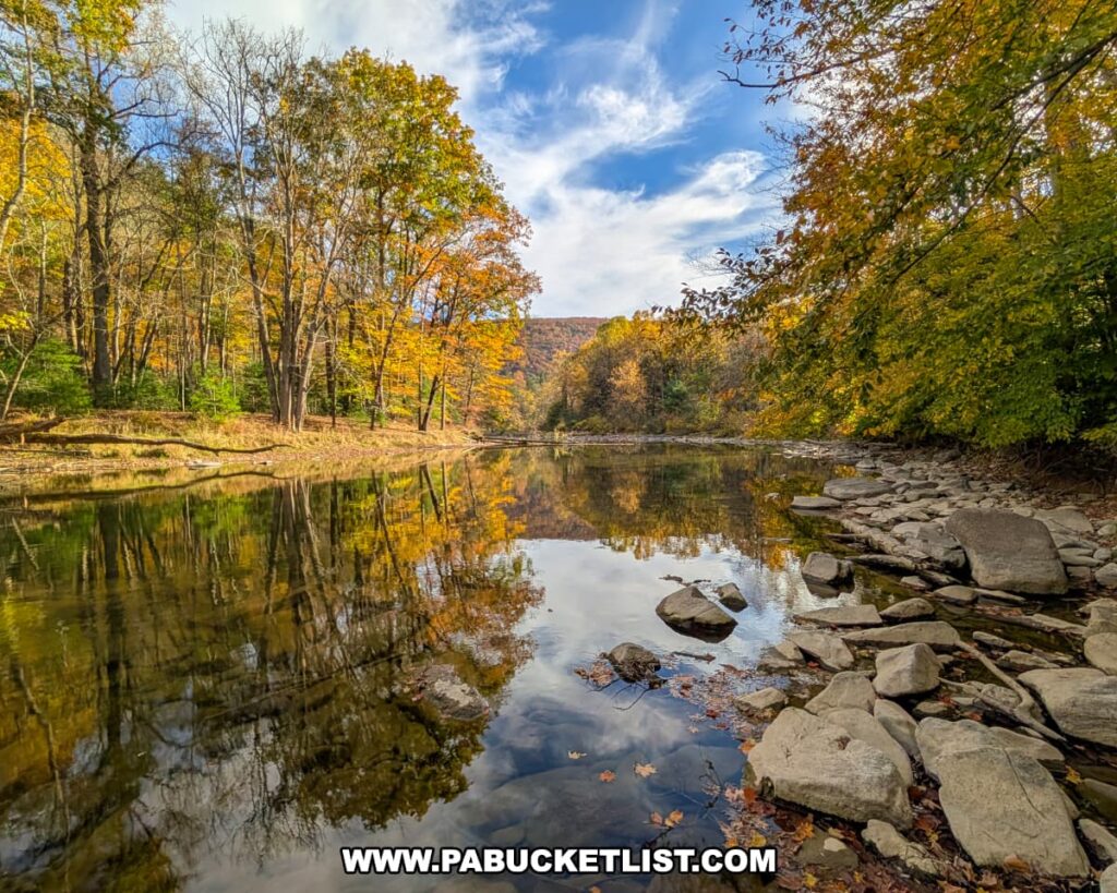 A calm stretch of Penn’s Creek reflects the surrounding autumn trees and blue sky near the Penn’s Creek Path in Bald Eagle State Forest, with smooth rocks lining the shore and forested hills visible in the distance.