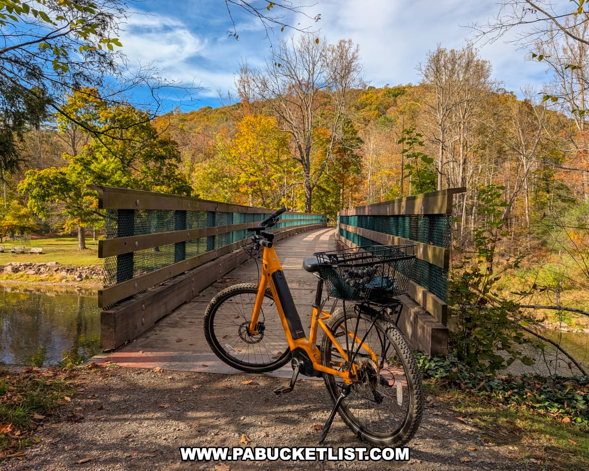 An orange bicycle with a black basket is parked at the entrance to a wooden bridge crossing Penn’s Creek along the Penn’s Creek Path in Bald Eagle State Forest, surrounded by colorful autumn foliage and forested hills under a partly cloudy sky.