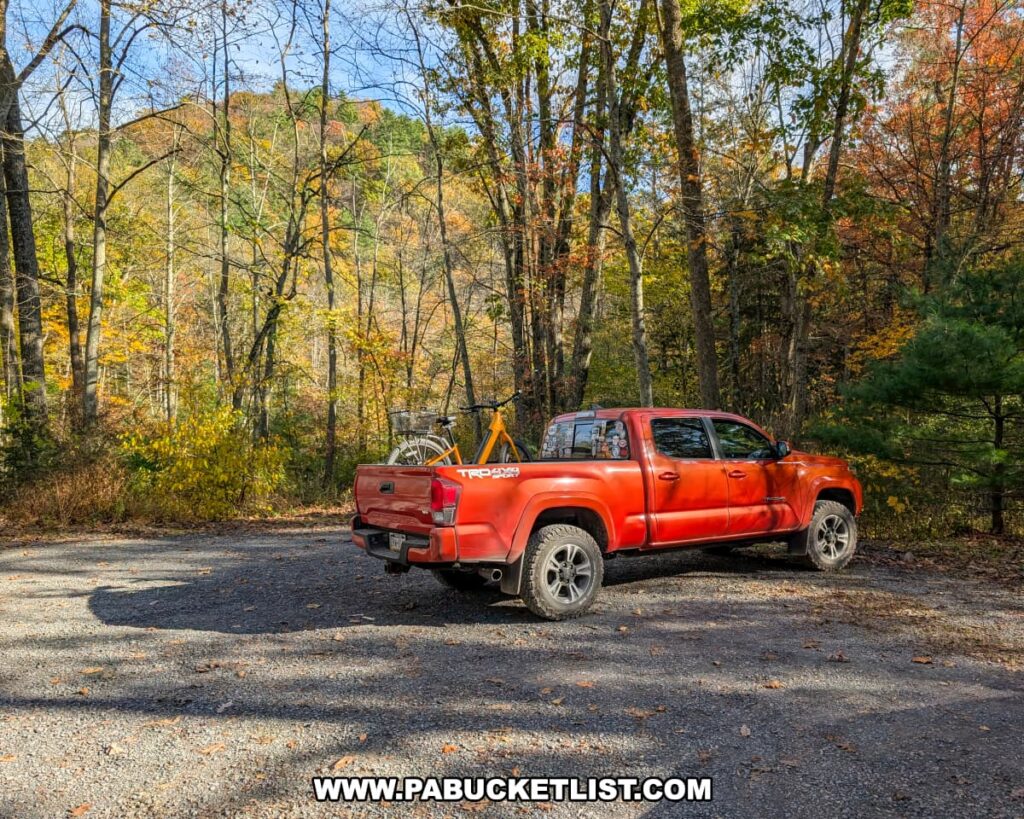 A red pickup truck with a bicycle in the bed is parked at a gravel lot near Poe Paddy State Park, surrounded by colorful autumn trees and forested hills along the Penn’s Creek Path in Bald Eagle State Forest.